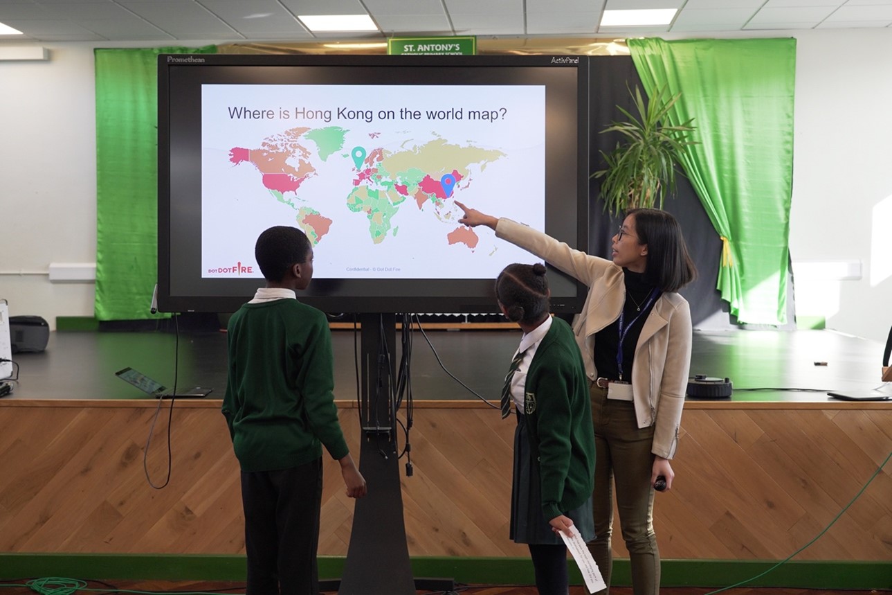 Dot Dot Fire's Hilary stands in front of a screen, pointing at the location of Hong Kong on a world map.